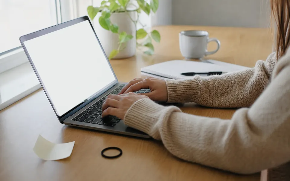 A bright home desk with an open laptop showing a plain white glow, a notebook and a mug